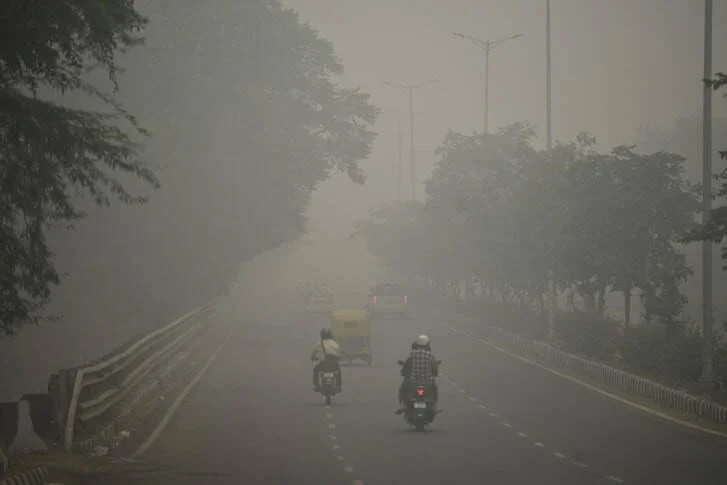 Dos motociclistas circulan por una avenida arbolada casi invisible bajo una densa capa de smog y contaminación atmosférica.