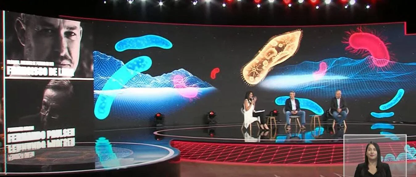 Conductora y dos panelistas en estudio de TV rodeados de animaciones de bacterias y microorganismos proyectadas en gran pantalla.