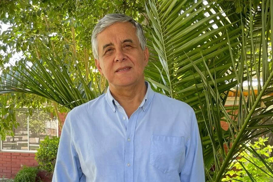 Hombre de cabello canoso con camisa celeste posa frente a palmeras y vegetación verde.