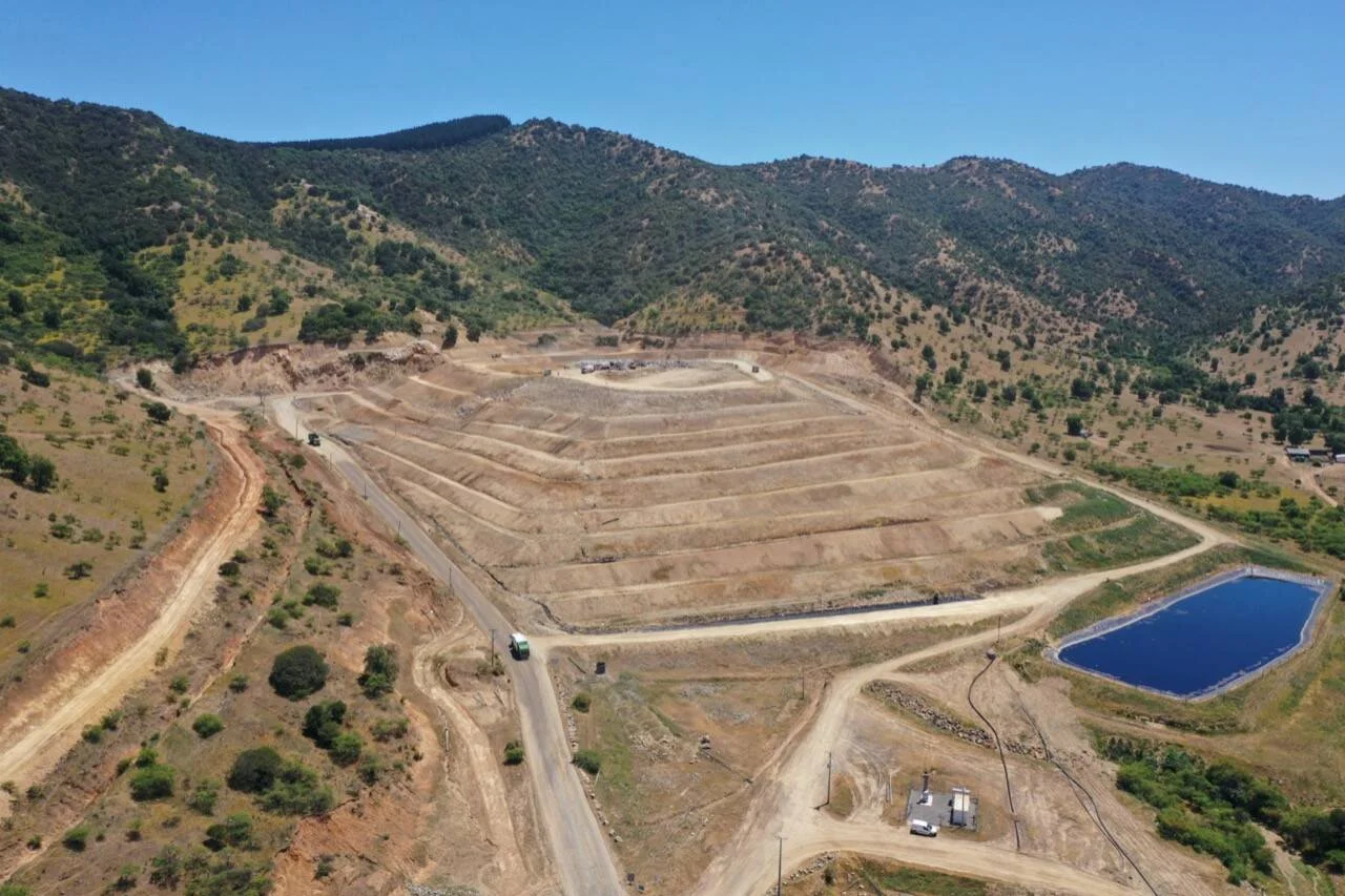 Vista aérea de un relleno sanitario con terrazas escalonadas, estanque de lixiviados y caminos de tierra rodeado de cerros vegetados.