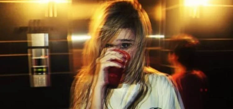 Joven rubia bebe de un vaso rojo en un entorno oscuro con luces amarillas, en aparente contexto de fiesta nocturna.