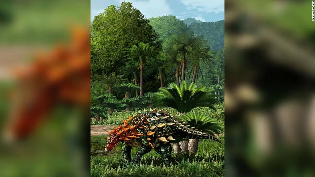 Recreación artística de un dinosaurio anquilosaurio acorazado con púas naranjas caminando entre helechos y palmeras prehistóricas.