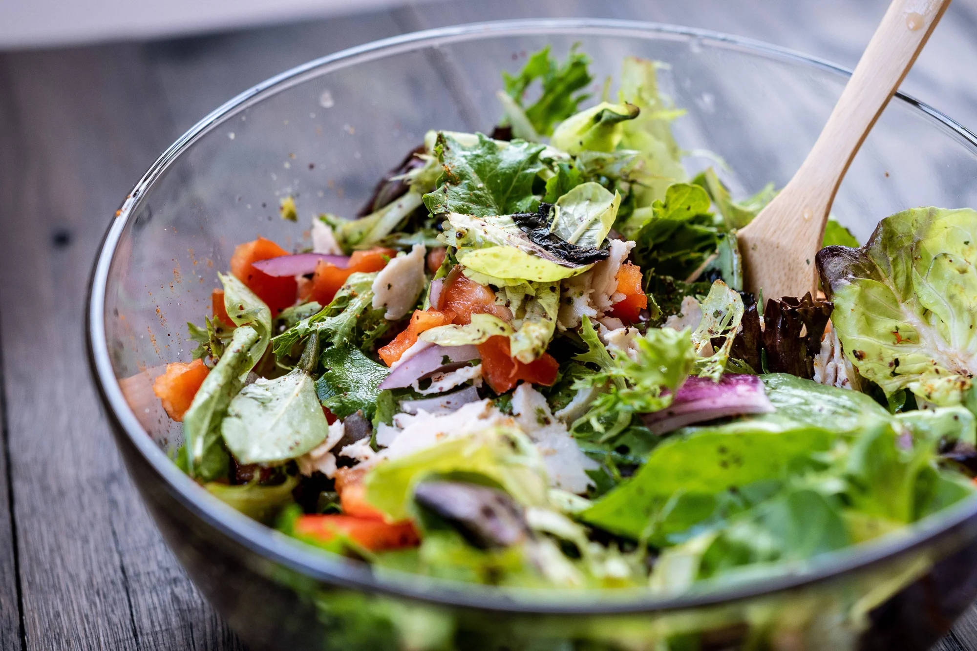 Ensalada de hojas verdes mixtas con tomate, cebolla morada y pollo en un tazón de vidrio con cuchara de madera.