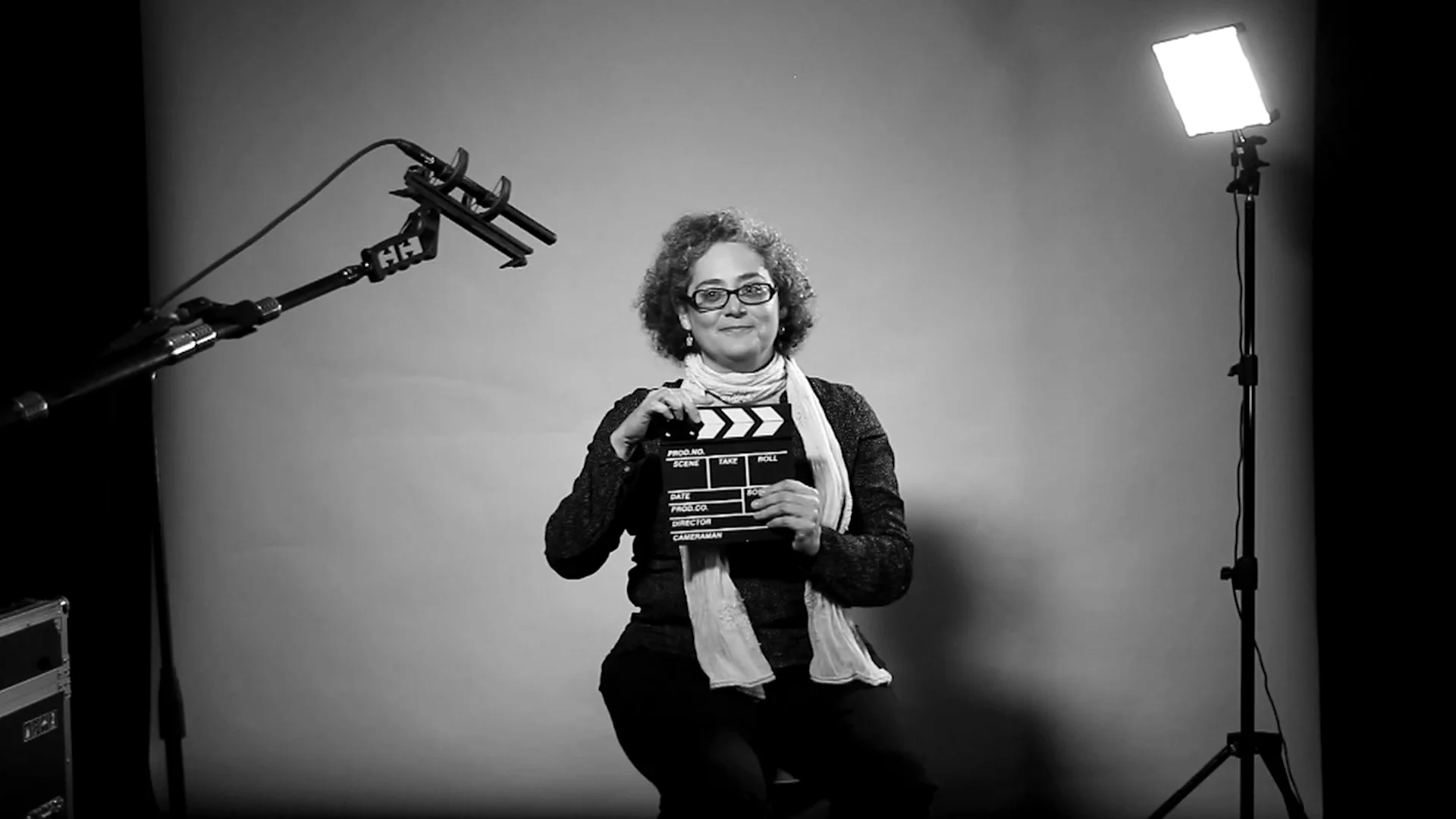 Mujer con gafas sostiene una claqueta de cine en un set profesional con micrófono boom y luz de estudio, fotografía en blanco y negro.