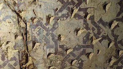 Mosaico arqueológico deteriorado con intrincados patrones geométricos en tonos ocres y morados sobre superficie de piedra.