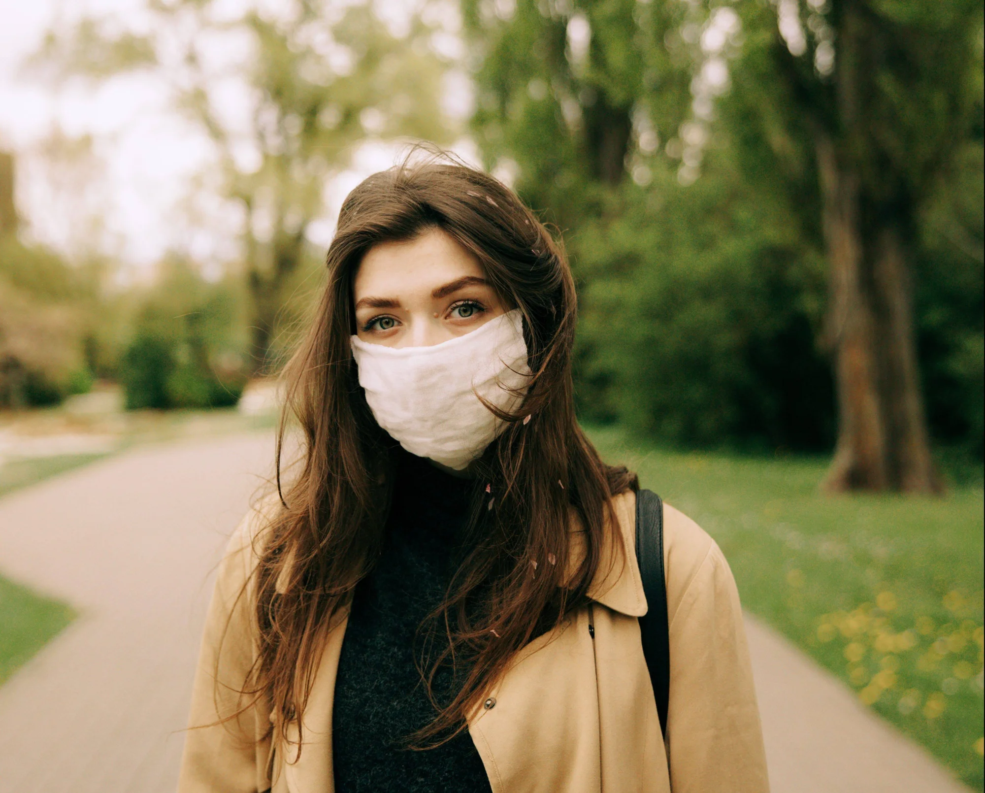 Mujer joven con mascarilla de tela blanca camina por un parque arbolado con abrigo beige.