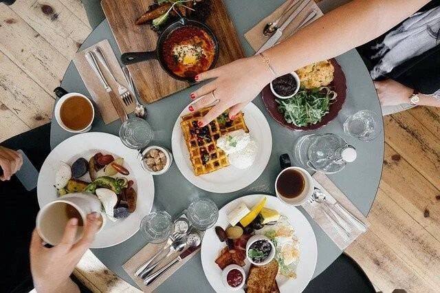 Vista aérea de una mesa de brunch con waffles, huevos, ensaladas, café y varios platos compartidos entre comensales.
