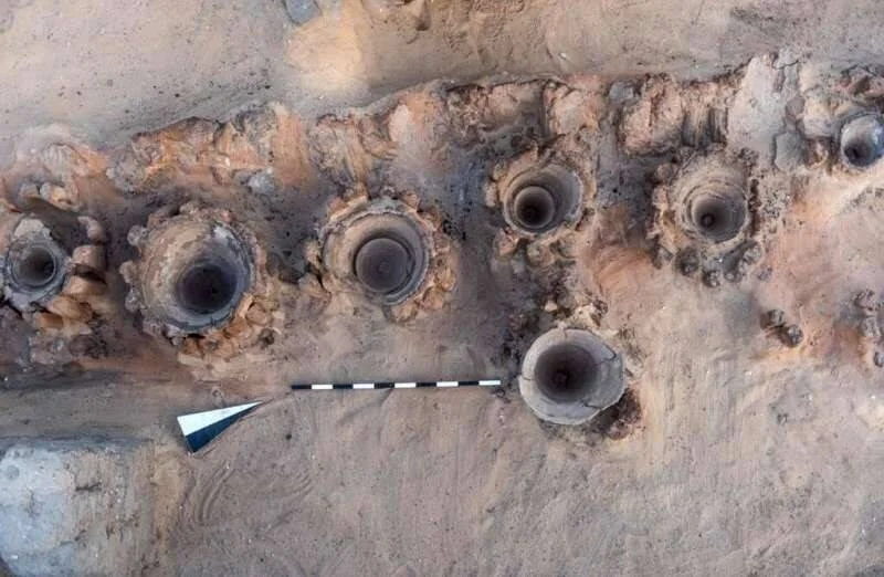 Vista aérea de vasijas cerámicas antiguas incrustadas en el suelo durante una excavación arqueológica, con escala métrica.
