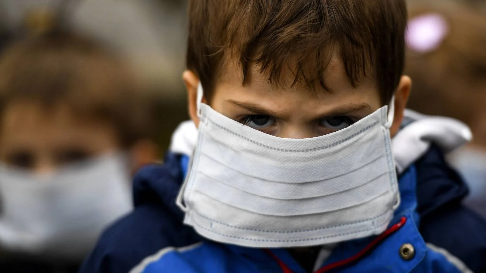 Niño con mascarilla quirúrgica blanca mirando a cámara, con otro menor con mascarilla al fondo.
