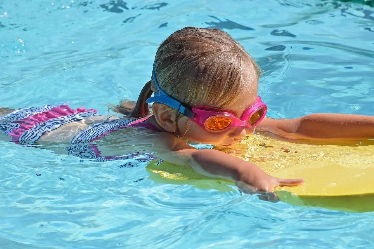 Niña con gafas de natación rosas practica natación apoyada en una tabla flotadora amarilla en una piscina.