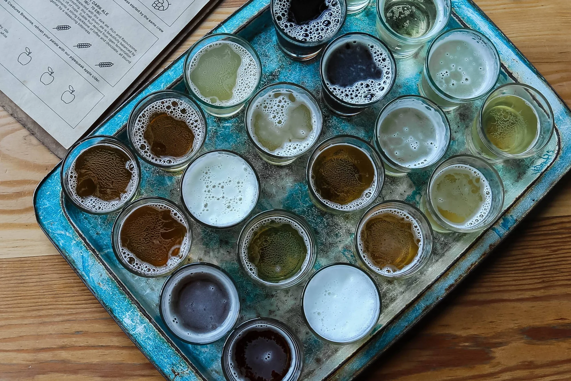 Bandeja metálica con múltiples vasos pequeños de cervezas artesanales de distintos colores y estilos, junto a un menú impreso.