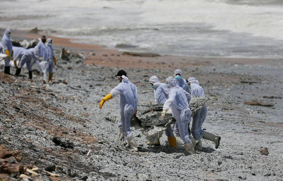 Trabajadores con trajes blancos de protección y guantes retiran residuos contaminantes de una playa afectada por un derrame.