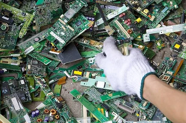 Una mano con guante blanco revuelve una gran acumulación de placas de circuito electrónico en un sitio de reciclaje de residuos electrónicos.