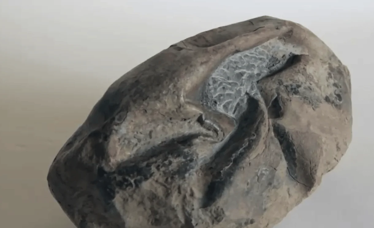 Escultura abstracta de un rostro humano modelado en arcilla o piedra de tonos grises y marrones.