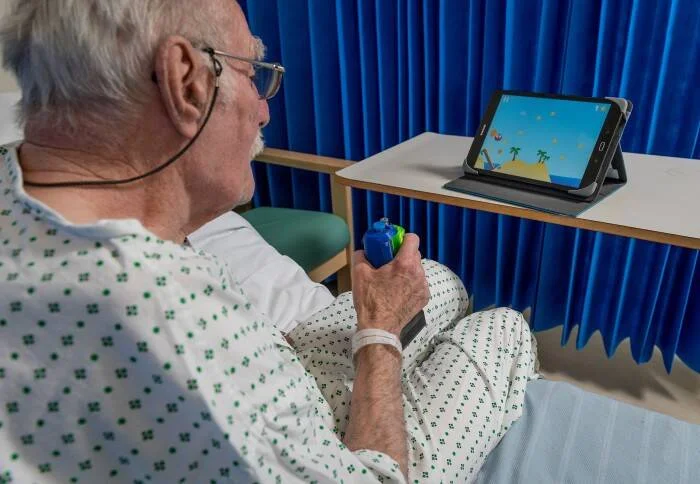 Adulto mayor hospitalizado sostiene un control y juega un videojuego en una tableta apoyada sobre una mesa.