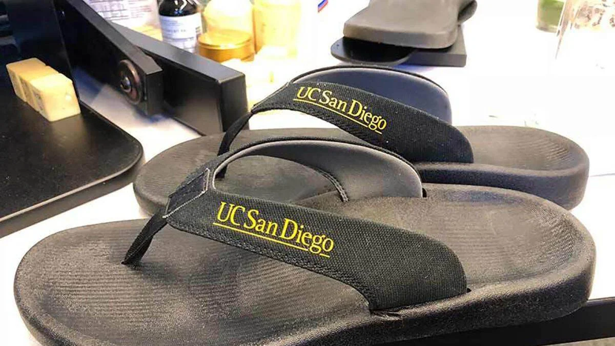 Par de sandalias tipo flip-flop negras con el logo 'UC San Diego' en letras doradas sobre las tiras.