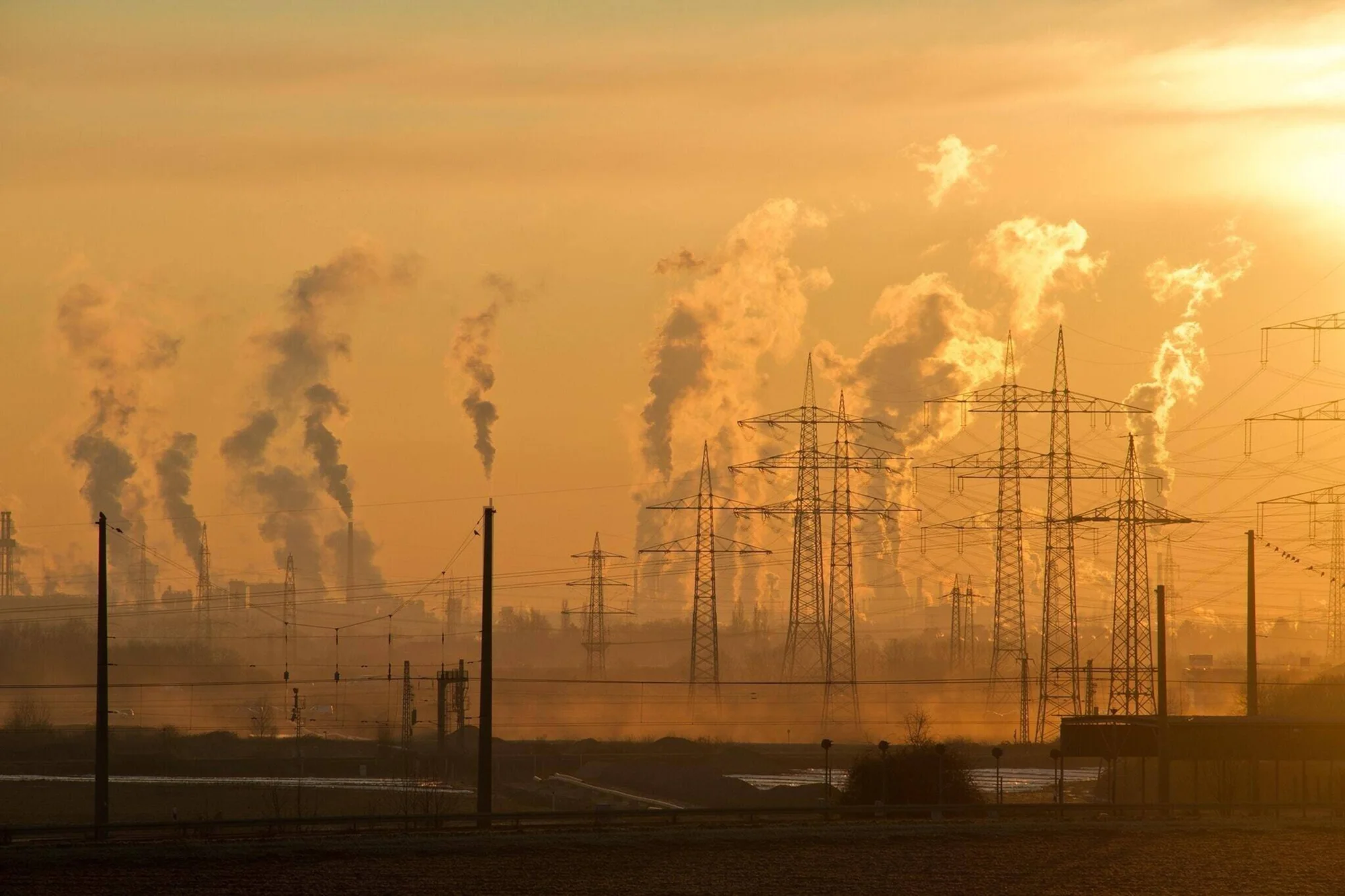 Zona industrial con múltiples chimeneas emitiendo humo y torres de alta tensión bajo un cielo anaranjado al atardecer.