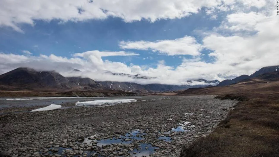 Lecho pedregoso de río ártico con fragmentos de hielo flotante, rodeado de montañas cubiertas de nubes y tundra.