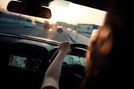 Persona conduciendo un auto en autopista con navegador GPS, vista desde el asiento trasero con luz de atardecer.