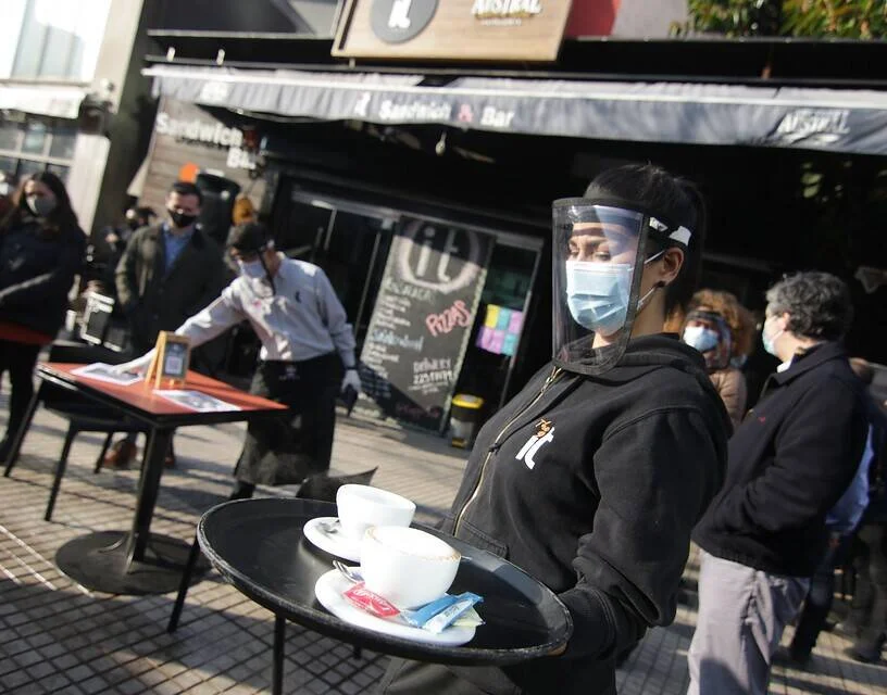 Moza con barbijo y escudo facial lleva una bandeja con tazas de café en la terraza de un bar durante la pandemia.