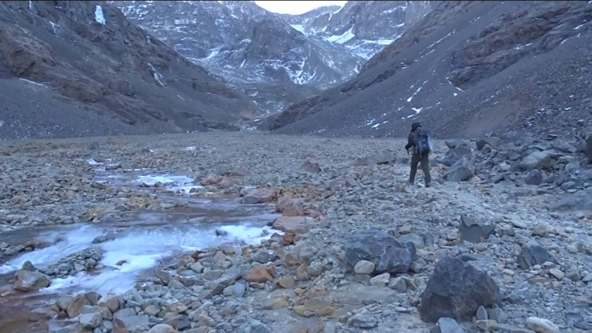 Una persona con mochila camina junto a un arroyo glaciar en un valle rocoso rodeado de montañas nevadas.