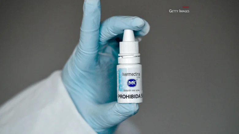 Mano con guante médico azul sostiene un frasco de ivermectina con la leyenda 'PROHIBIDA' en su etiqueta.