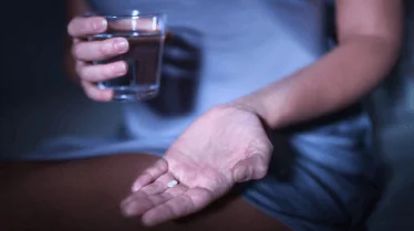 Mano extendida sosteniendo una pastilla blanca mientras la otra mano sujeta un vaso de agua.