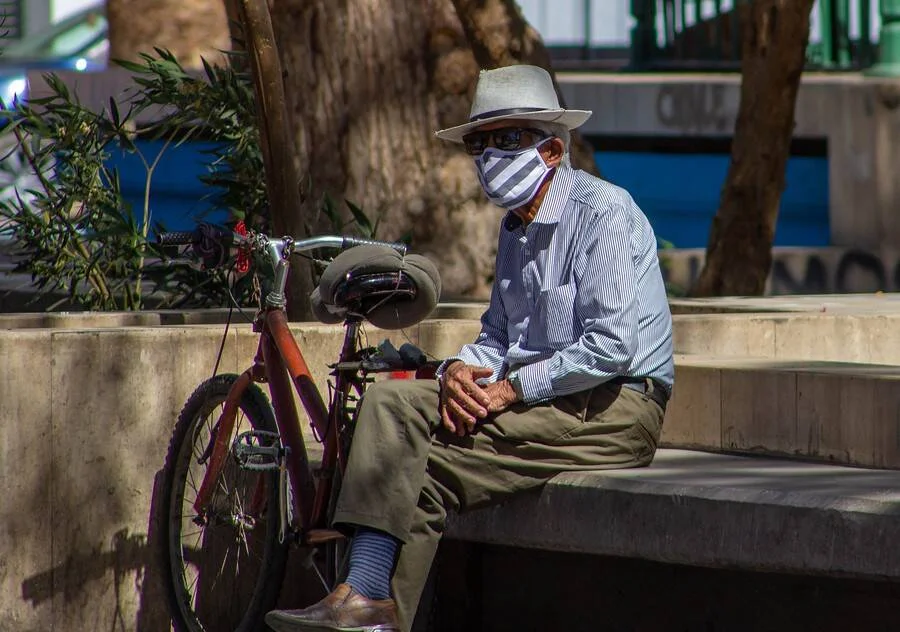 Hombre mayor con sombrero y barbijo descansa en un banco de plaza junto a su bicicleta.