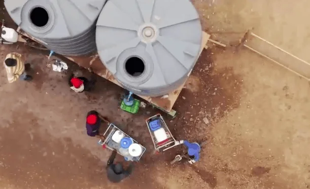 Vista desde dron de personas cargando bidones de agua junto a grandes tanques plásticos en terreno árido.