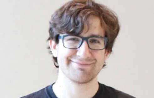 Hombre joven de cabello rizado, anteojos de marco oscuro y remera negra, sonriendo frente a un fondo claro.