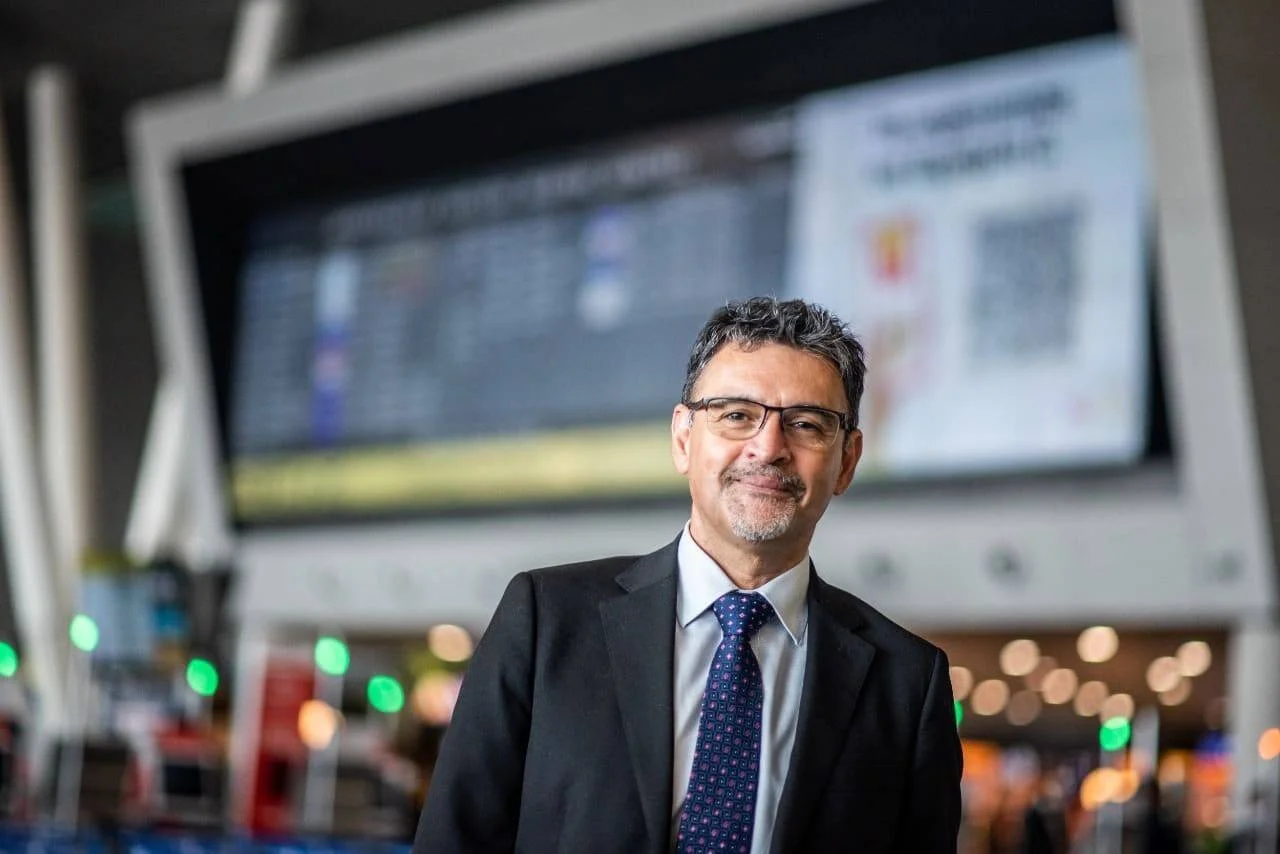 Hombre con traje oscuro y corbata azul posa sonriente frente a pantallas digitales en una terminal de aeropuerto.