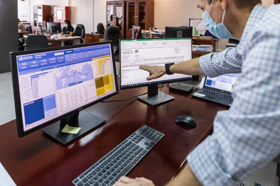 Funcionario con mascarilla señala una pantalla con el panel de monitoreo de salud de la OMS Dubai Hub en una oficina.