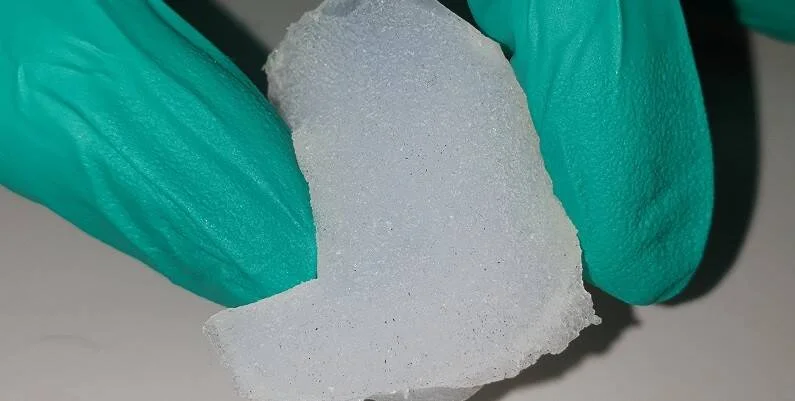 Manos con guantes verdes sostienen una lámina de material poroso translúcido de aspecto granulado.