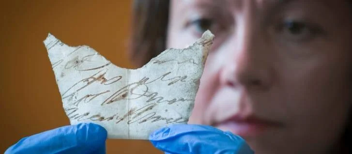 Una persona con guantes azules sostiene un pequeño fragmento de pergamino antiguo con escritura manuscrita en tinta marrón.