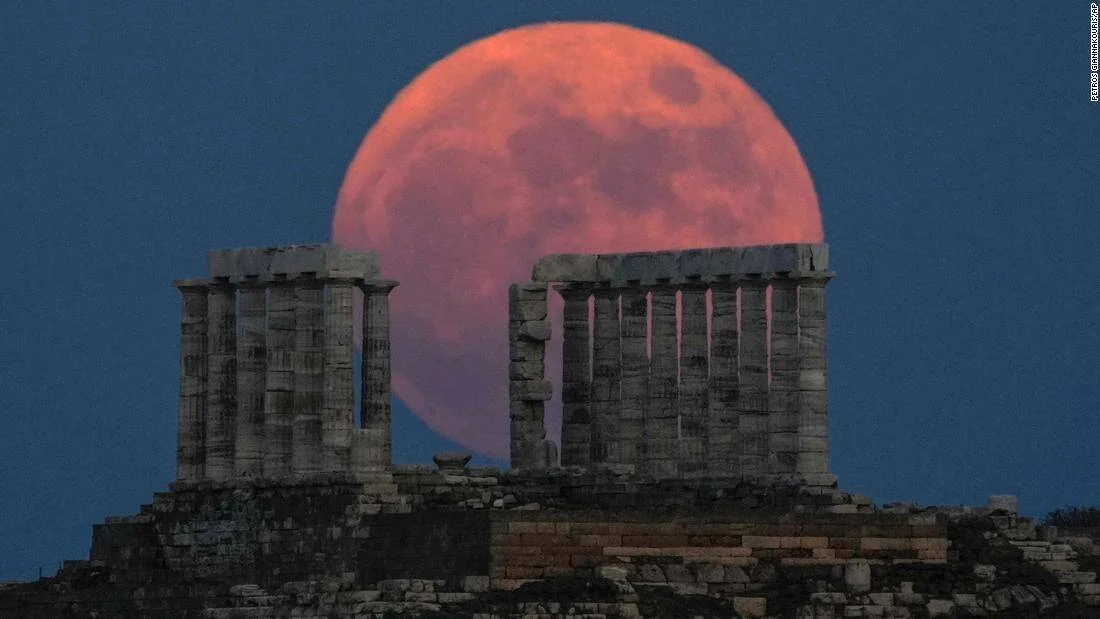 Luna llena de color rojizo se eleva detrás de las ruinas de un templo griego con columnas sobre una colina al anochecer.