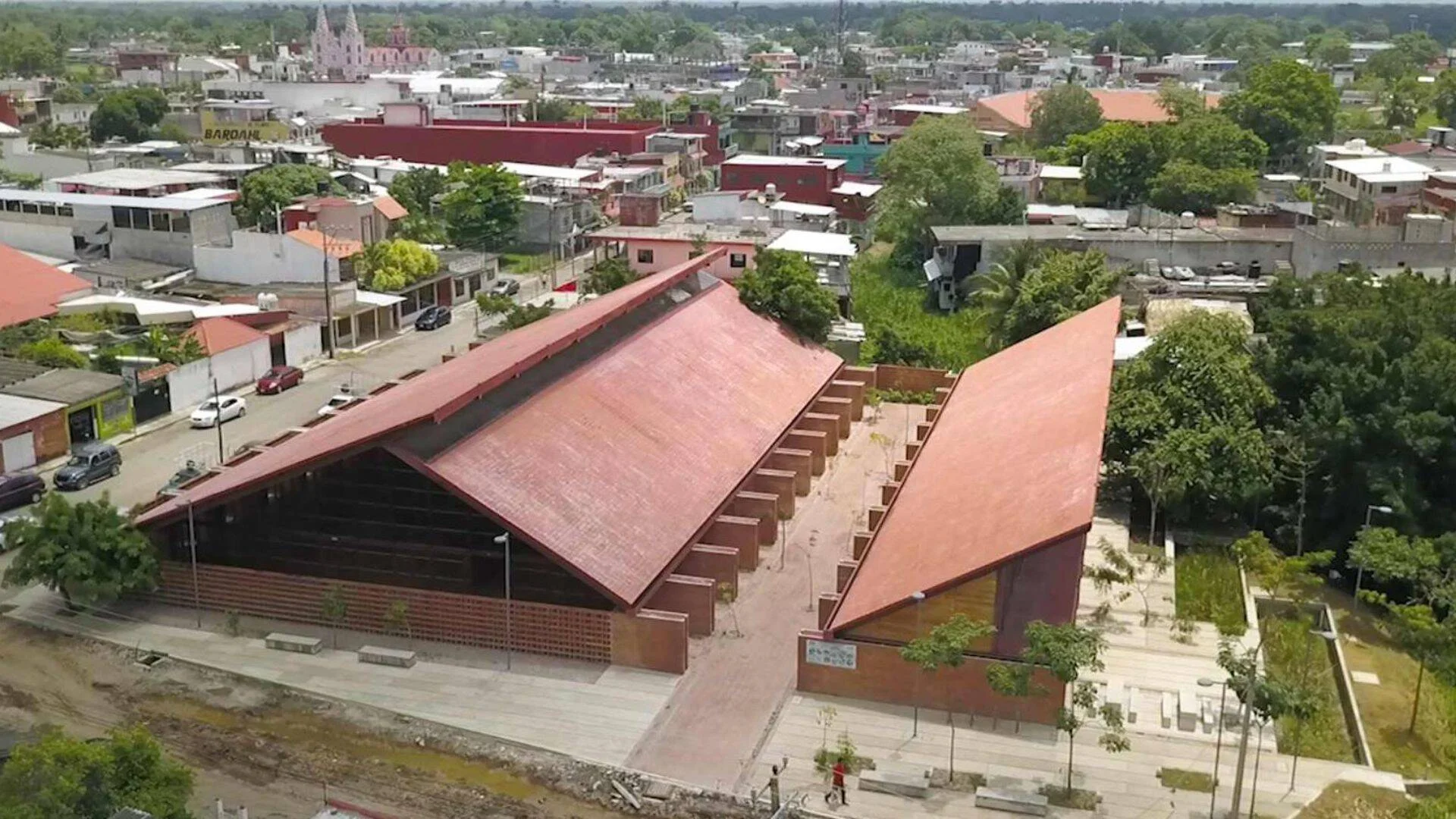 Vista aérea de un edificio de gran cubierta inclinada color rojo ladrillo rodeado de una ciudad con vegetación y una iglesia al fondo.