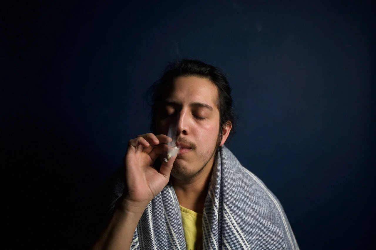Hombre joven con cabello recogido y manta al hombro fuma un cigarrillo con los ojos cerrados ante fondo oscuro.