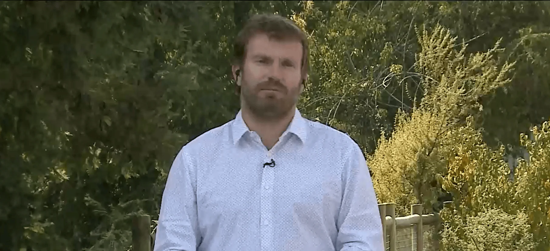 Hombre con camisa celeste y micrófono de solapa frente a cámara en un jardín con árboles de fondo.