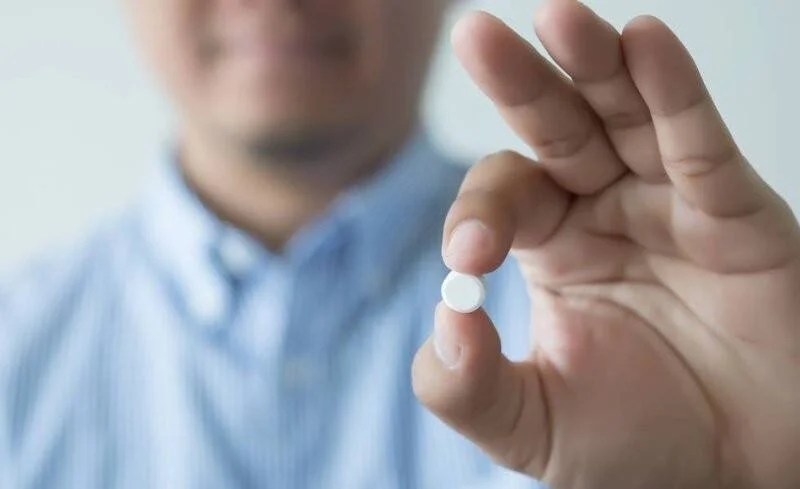 Una persona con camisa azul sostiene una pequeña pastilla blanca entre sus dedos índice y pulgar.