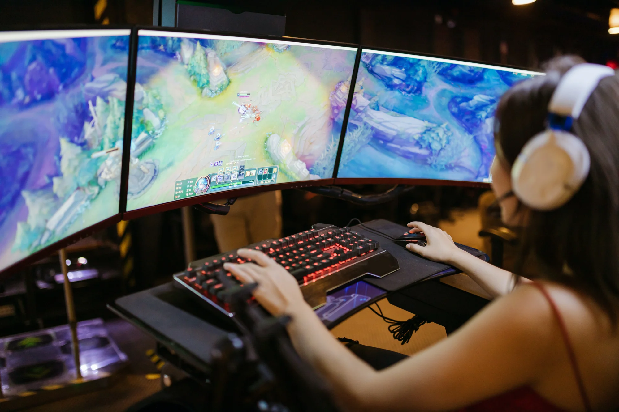 Mujer con auriculares juega un videojuego de estrategia en una configuración de triple monitor con teclado mecánico iluminado.