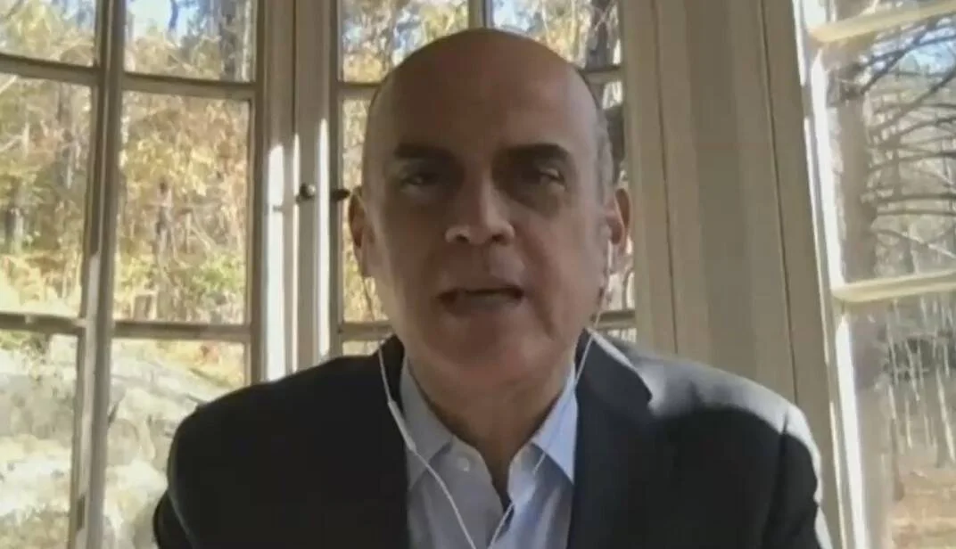 Hombre calvo con saco oscuro participa en una videollamada desde una habitación con ventanas y árboles al fondo.