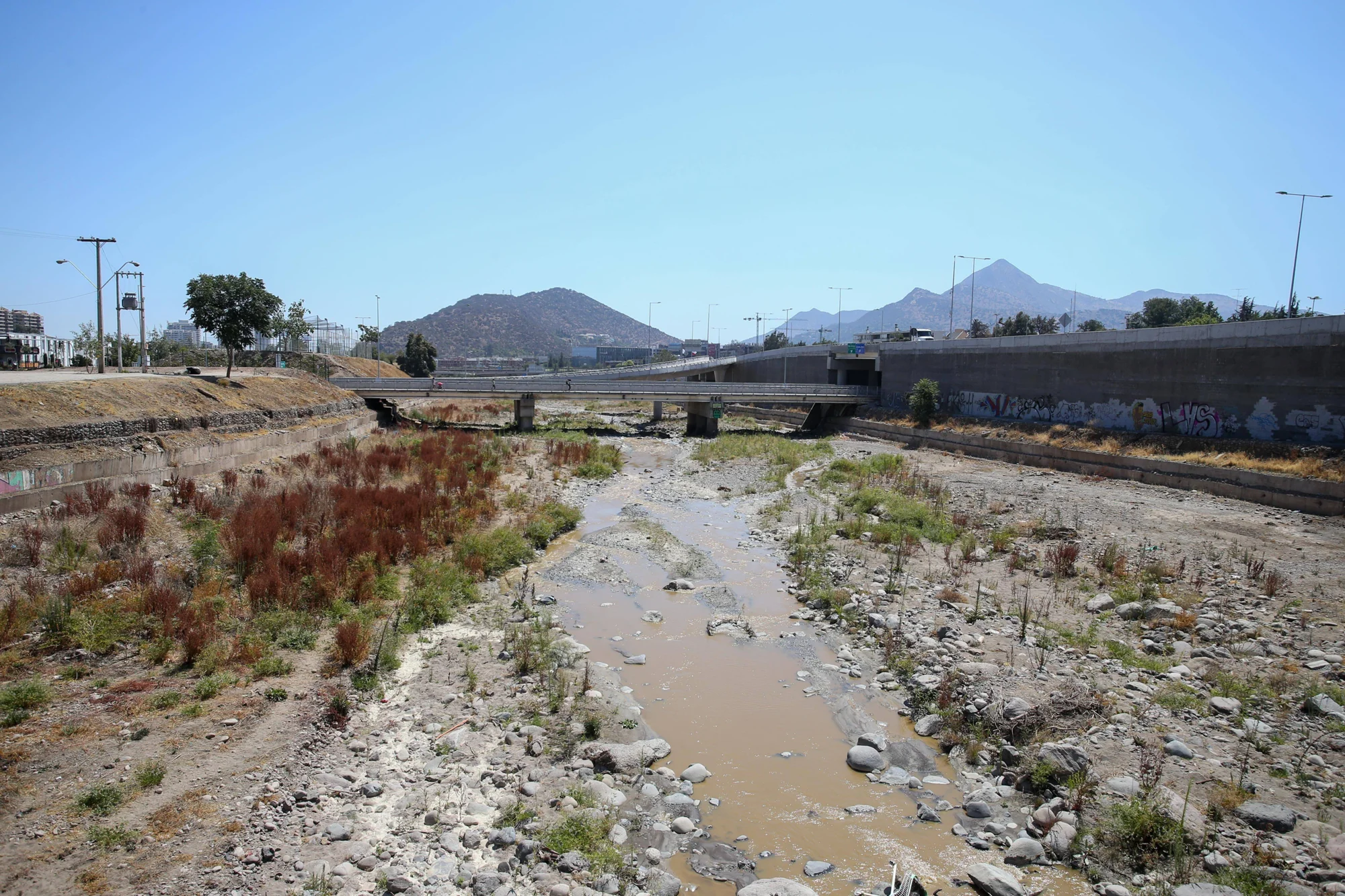 Cauce de río casi seco con escaso caudal lodoso, rodeado de piedras y vegetación seca, con un puente urbano y cerros al fondo.