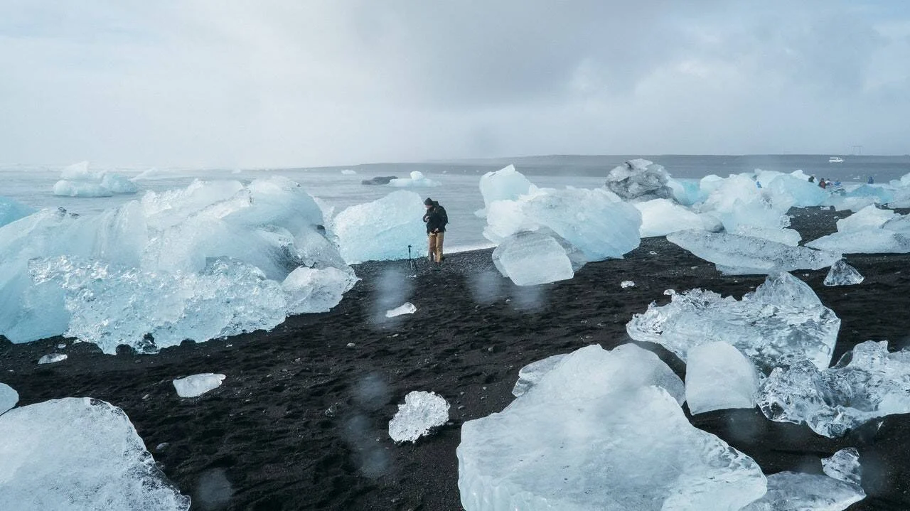 Una persona fotografía bloques de hielo azul turquesa dispersos sobre una playa de arena negra volcánica bajo un cielo nublado.