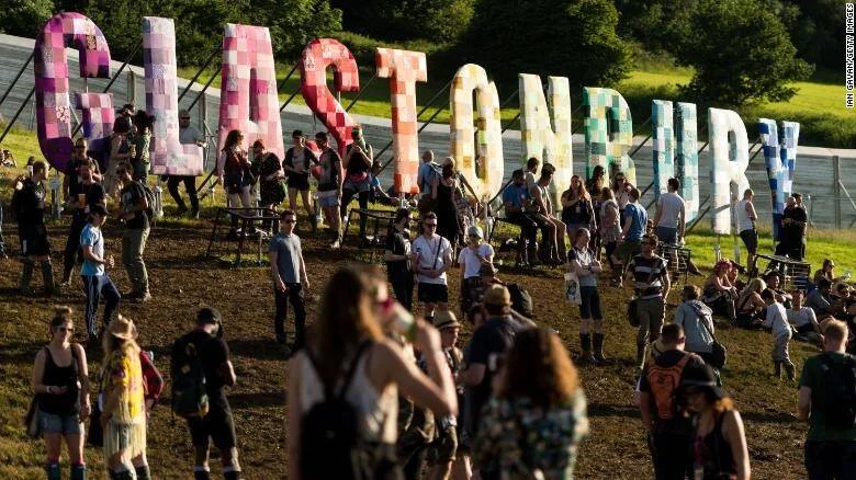 Multitud de personas reunidas frente al cartel gigante colorido del Festival de Glastonbury en un campo verde.
