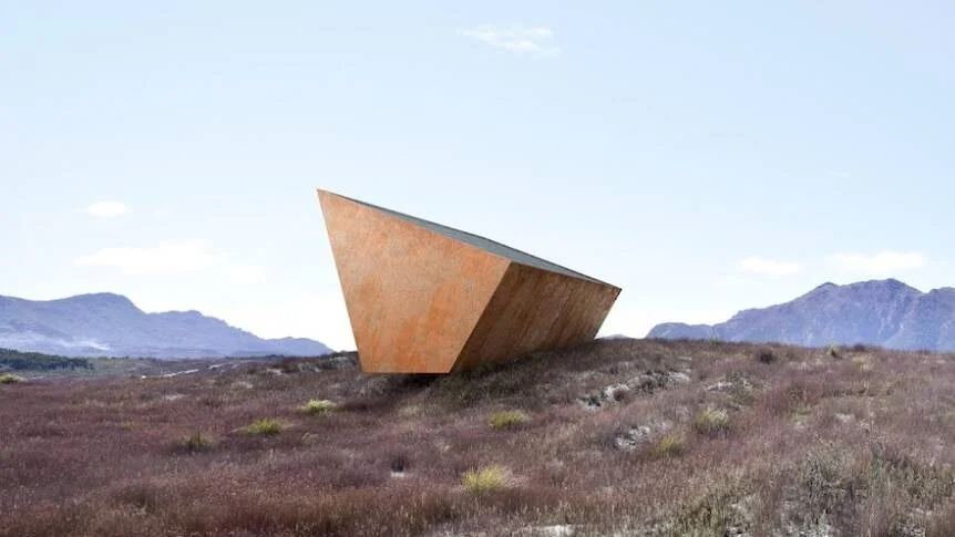 Edificio de forma triangular angular con revestimiento de acero oxidado, sobre una colina con vegetación árida y montañas al fondo.