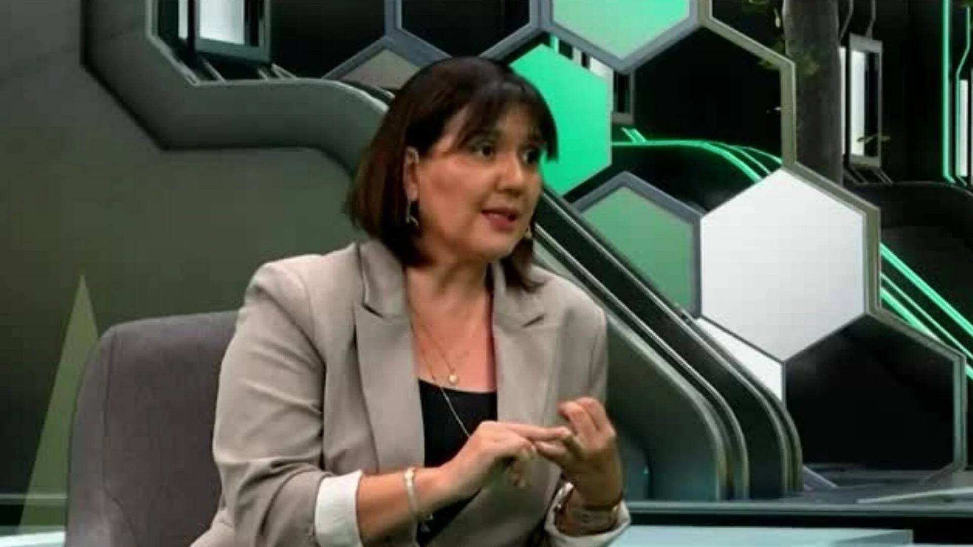 Mujer con blazer gris gesticula mientras habla sentada en un estudio de televisión con decorado hexagonal verde y negro.