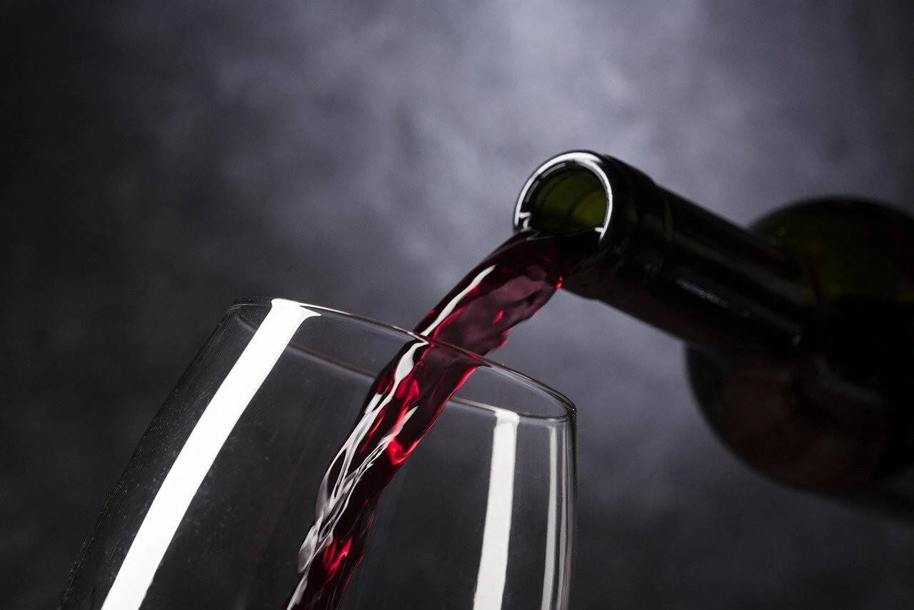 Vino tinto siendo vertido desde una botella de vidrio verde a una copa transparente sobre fondo gris oscuro.