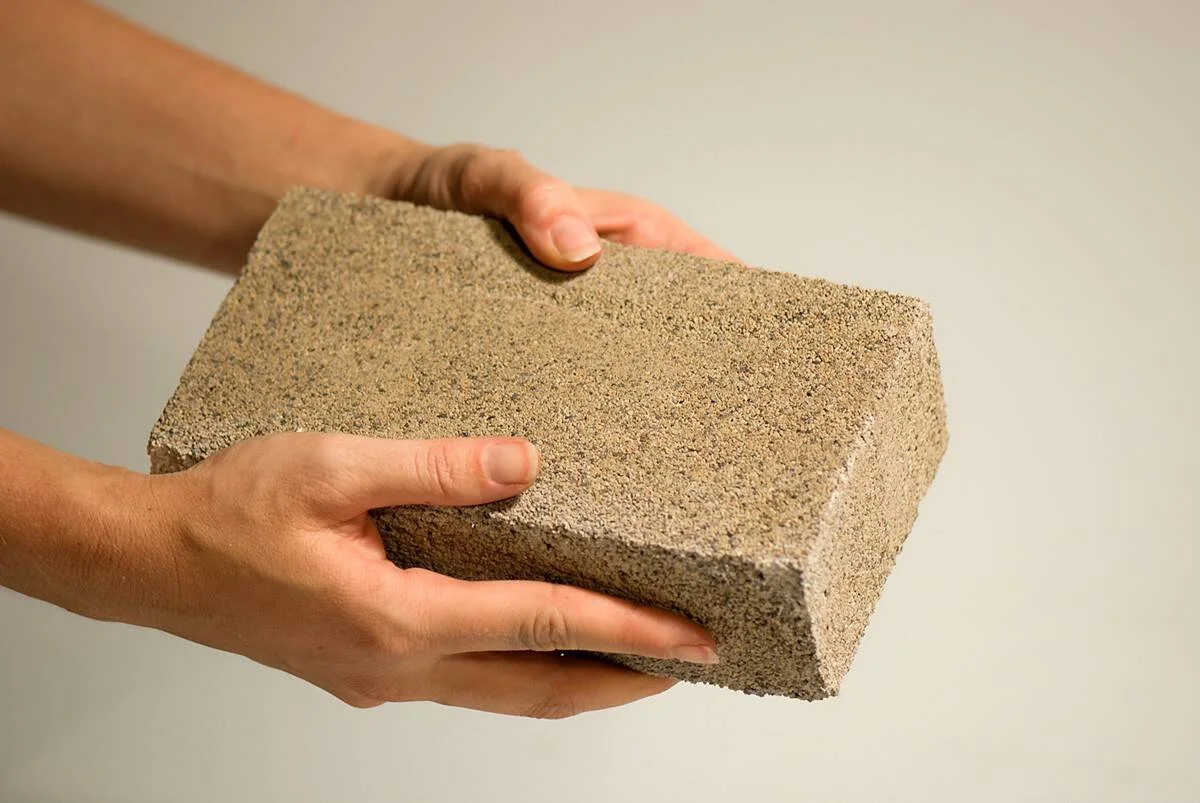 Dos manos sostienen un ladrillo de textura arenosa de color beige grisáceo sobre fondo claro.