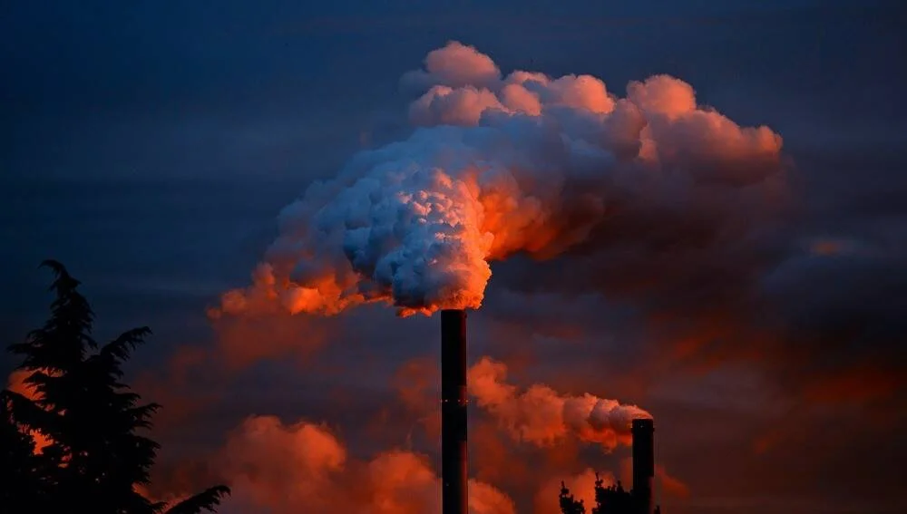 Chimeneas de una planta industrial expulsan grandes columnas de humo iluminadas por la luz rojiza del atardecer.