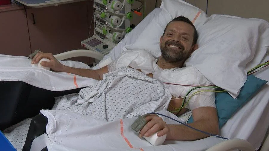 Hombre con bata hospitalaria sonríe desde su cama, conectado a monitores y cables médicos.